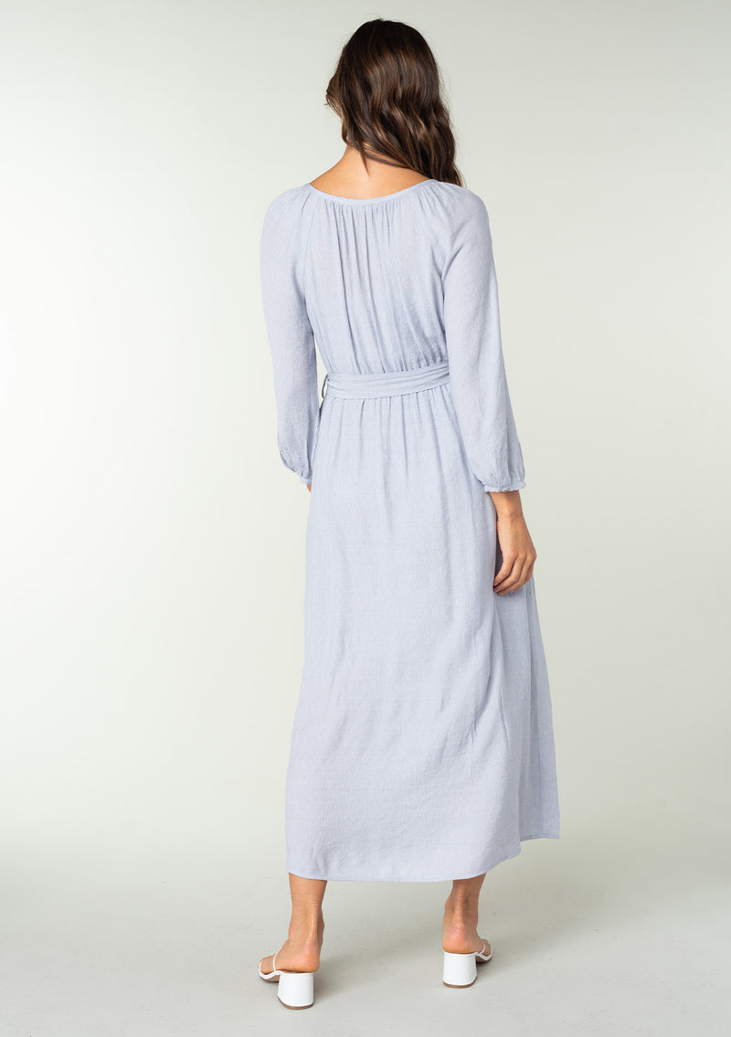 [Color: Dusty Lilac] A back facing image of a brunette model wearing a classic light purple bohemian maxi dress. With long sleeves, a round neckline, a button front, side pockets, and a self tie waist belt. 