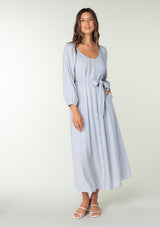 [Color: Dusty Lilac] A full body front facing image of a brunette model wearing a classic light purple bohemian maxi dress. With long sleeves, a round neckline, a button front, side pockets, and a self tie waist belt. 