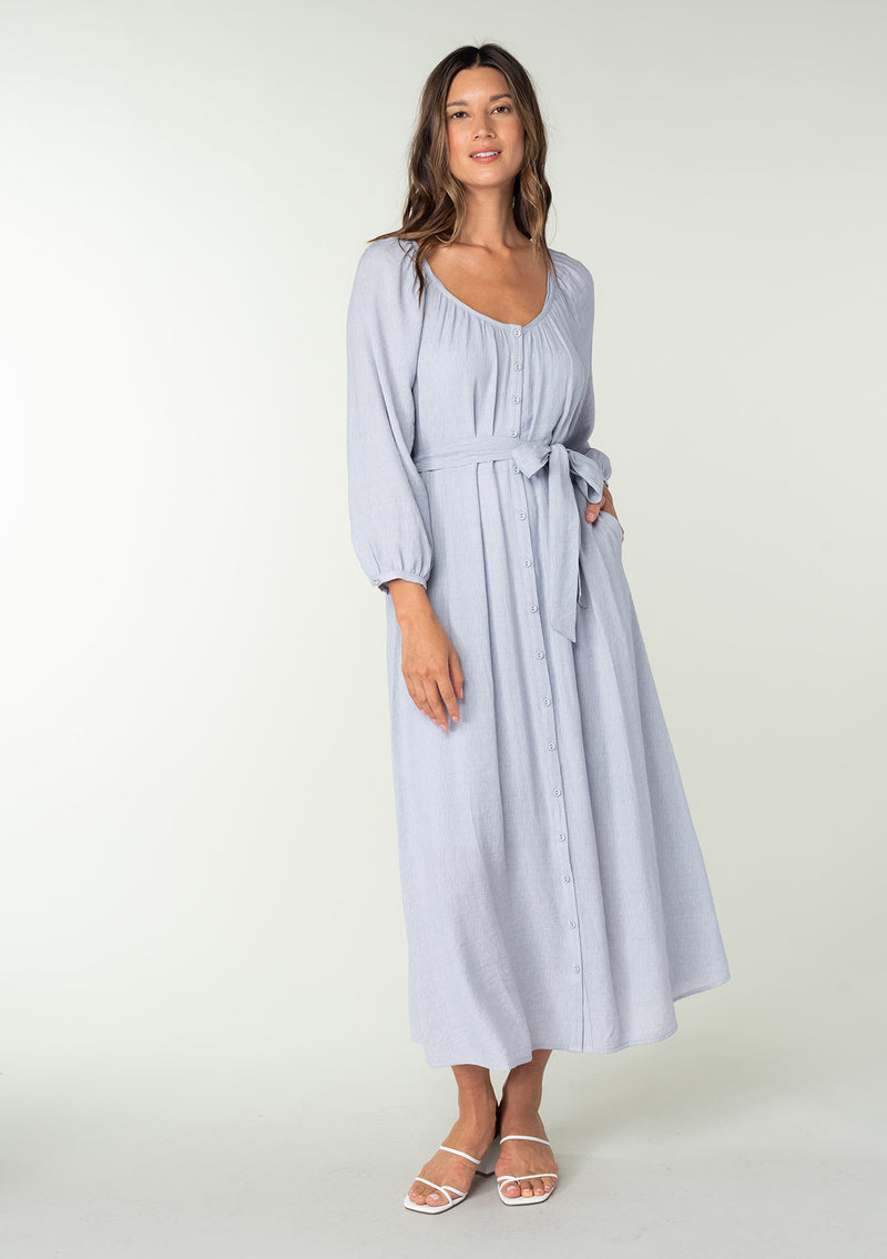 [Color: Dusty Lilac] A full body front facing image of a brunette model wearing a classic light purple bohemian maxi dress. With long sleeves, a round neckline, a button front, side pockets, and a self tie waist belt. 