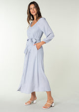 [Color: Dusty Lilac] A side facing image of a brunette model wearing a classic light purple bohemian maxi dress. With long sleeves, a round neckline, a button front, side pockets, and a self tie waist belt. 