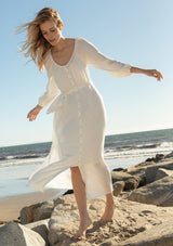[Color: White] A full body front facing image of a blonde model at the beach wearing a classic white bohemian maxi dress. With long sleeves, a round neckline, a button front, side pockets, and a self tie waist belt.