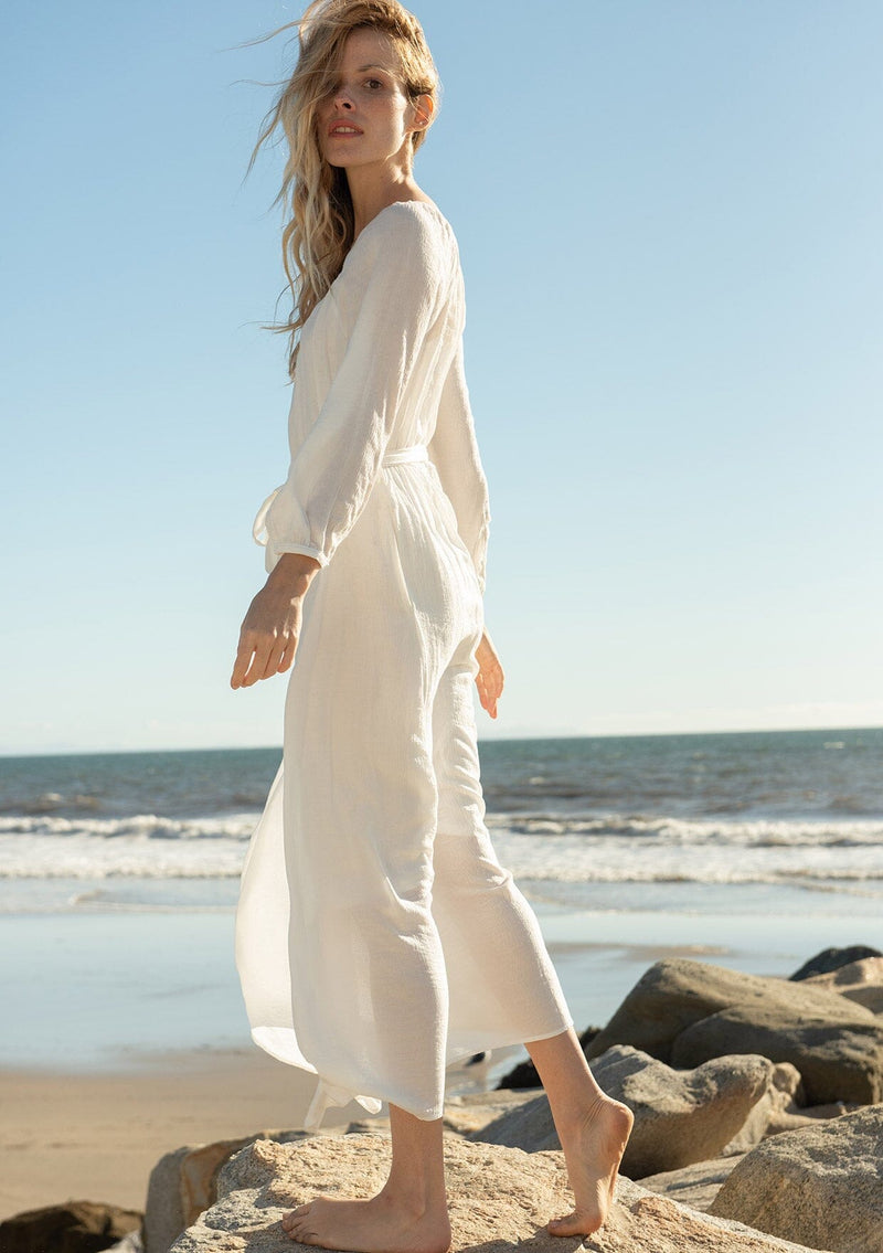 [Color: White] A side facing image of a blonde model at the beach wearing a classic white bohemian maxi dress. With long sleeves, a round neckline, a button front, side pockets, and a self tie waist belt.