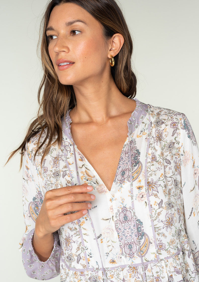 [Color: Natural/Dusty Lilac] A close up front facing image of a brunette model wearing a light lilac purple and natural off white mixed floral print mini dress. With an ultra flowy baby doll dress silhouette, long sleeves, a tiered skirt, and a split neckline with ties. 