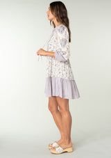 [Color: Natural/Dusty Lilac] A side facing image of a brunette model wearing a light lilac purple and natural off white mixed floral print mini dress. With an ultra flowy baby doll dress silhouette, long sleeves, a tiered skirt, and a split neckline with ties. 