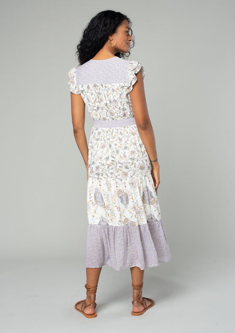 [Color: Natural/Dusty Lilac] A back facing image of a brunette model wearing a bohemian mid length dress in an off white and light purple mixed floral print. With short flutter cap sleeves, a smocked elastic waist, a tiered skirt, and a v neckline.