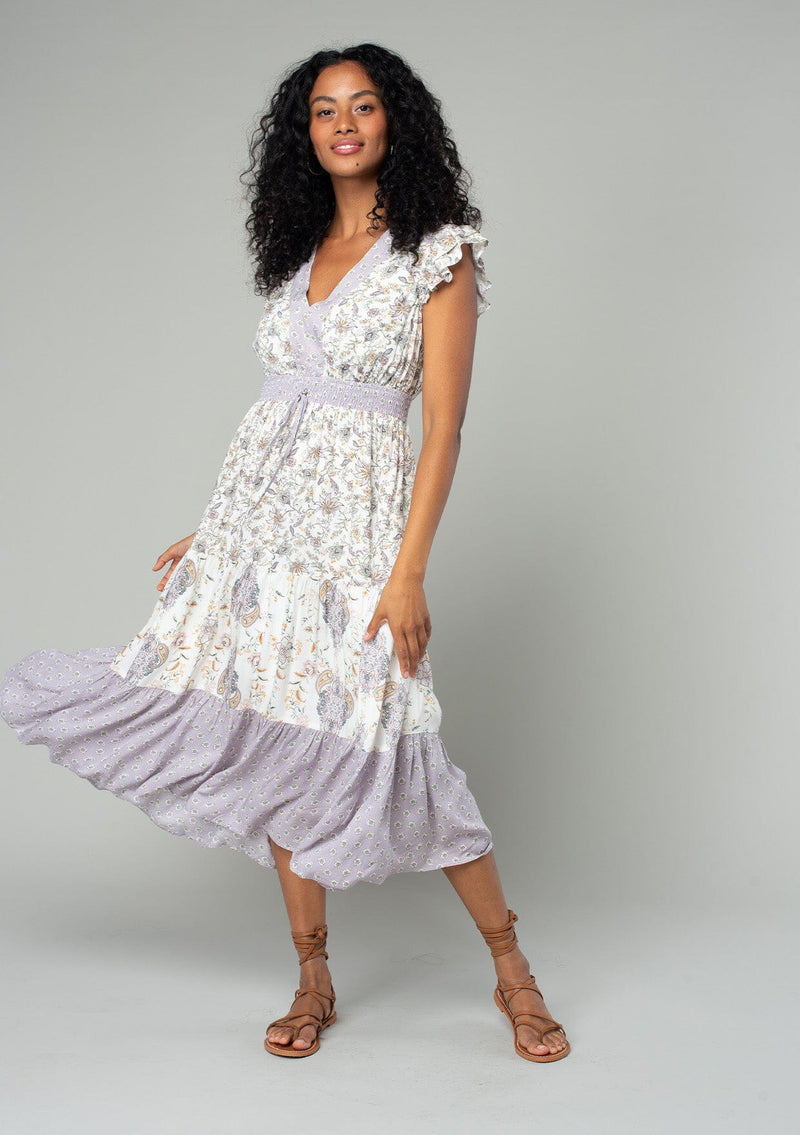 [Color: Natural/Dusty Lilac] A front facing image of a brunette model wearing a bohemian mid length dress in an off white and light purple mixed floral print. With short flutter cap sleeves, a smocked elastic waist, a tiered skirt, and a v neckline.