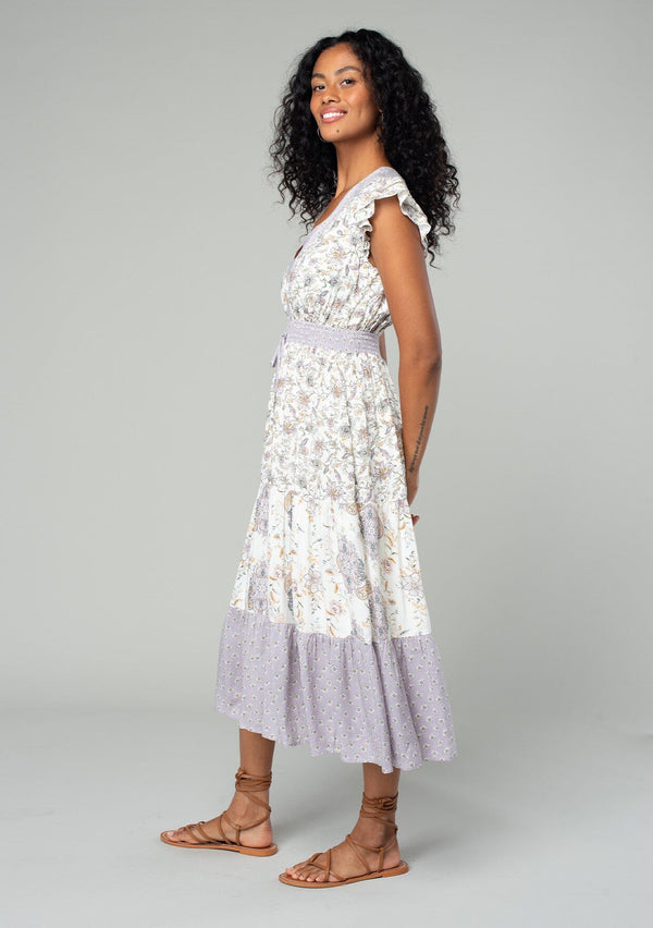 [Color: Natural/Dusty Lilac] A side facing image of a brunette model wearing a bohemian mid length dress in an off white and light purple mixed floral print. With short flutter cap sleeves, a smocked elastic waist, a tiered skirt, and a v neckline. 