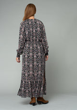 [Color: Black/Rose] A back facing image of a red headed model wearing a classic bohemian maxi dress in a black and pink paisley print. With long sleeves, side slits, and an elastic waist. 