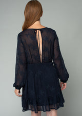 [Color: Navy] A back facing image of a red headed model wearing a navy blue embroidered chiffon mini dress. Perfect for the holidays or weddings, featuring long sleeves, a tiered skirt, an open back keyhole with tie closure, and a smocked elastic waist. 