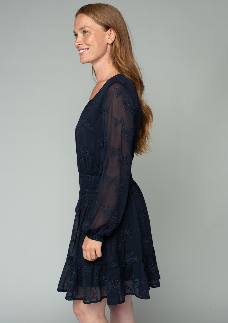 [Color: Navy] A side facing image of a red headed model wearing a navy blue embroidered chiffon mini dress. Perfect for the holidays or weddings, featuring long sleeves, a tiered skirt, an open back keyhole with tie closure, and a smocked elastic waist. 