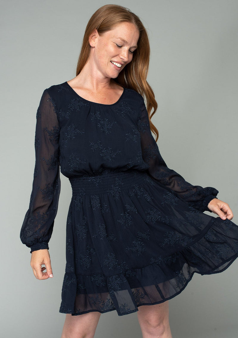 [Color: Navy] A half body front facing image of a red headed model wearing a navy blue embroidered chiffon mini dress. Perfect for the holidays or weddings, featuring long sleeves, a tiered skirt, an open back keyhole with tie closure, and a smocked elastic waist. 