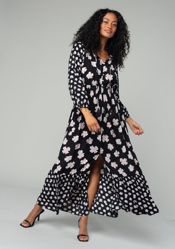 [Color: Black/Taupe] A front facing image of a brunette model wearing a classic bohemian maxi dress in a black and taupe mixed floral print. With long sleeves, a smocked elastic waist, a tiered flowy skirt, a button front top, and a split v neckline with tassel ties. 