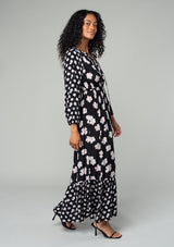 [Color: Black/Taupe] A side facing image of a brunette model wearing a classic bohemian maxi dress in a black and taupe mixed floral print. With long sleeves, a smocked elastic waist, a tiered flowy skirt, a button front top, and a split v neckline with tassel ties. 