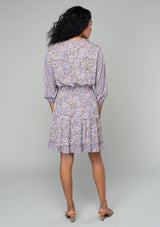 [Color: Grey/Natural] A back facing image of a brunette model wearing a classic bohemian mini dress in a grey and purple mixed floral print. With three quarter length sleeves, a split v neckline with tassel ties, a smocked elastic waist, and a ruffle trimmed tiered skirt. 