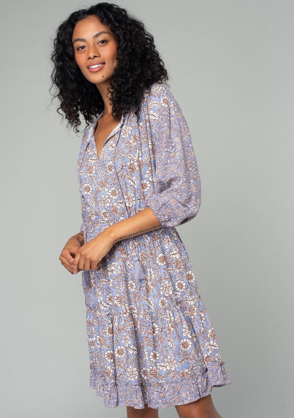 [Color: Grey/Natural] A half body side facing image of a brunette model wearing a classic bohemian mini dress in a grey and purple mixed floral print. With three quarter length sleeves, a split v neckline with tassel ties, a smocked elastic waist, and a ruffle trimmed tiered skirt. 