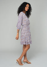 [Color: Grey/Natural] A full body side facing image of a brunette model wearing a classic bohemian mini dress in a grey and purple mixed floral print. With three quarter length sleeves, a split v neckline with tassel ties, a smocked elastic waist, and a ruffle trimmed tiered skirt. 