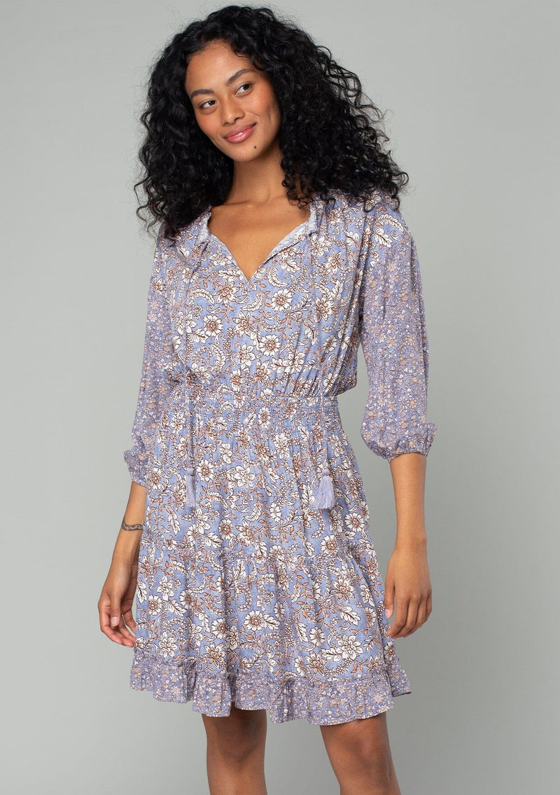 [Color: Grey/Natural] A half body front facing image of a brunette model wearing a classic bohemian mini dress in a grey and purple mixed floral print. With three quarter length sleeves, a split v neckline with tassel ties, a smocked elastic waist, and a ruffle trimmed tiered skirt. 