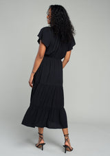 [Color: Black] A back facing image of a brunette model wearing a black bohemian spring mid length dress with short flutter sleeves, a tiered flowy skirt, an elastic waist, and lace trim detail at the top. 