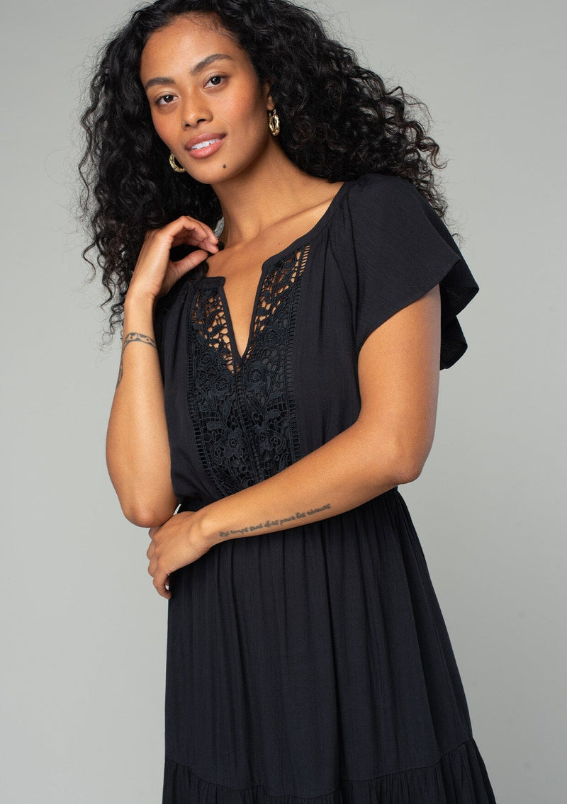 [Color: Black] A close up front facing image of a brunette model wearing a black bohemian spring mid length dress with short flutter sleeves, a tiered flowy skirt, an elastic waist, and lace trim detail at the top. 
