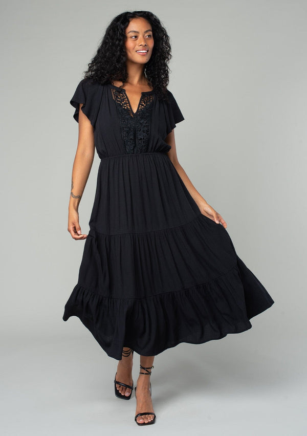 [Color: Black] A front facing image of a brunette model wearing a black bohemian spring mid length dress with short flutter sleeves, a tiered flowy skirt, an elastic waist, and lace trim detail at the top. 