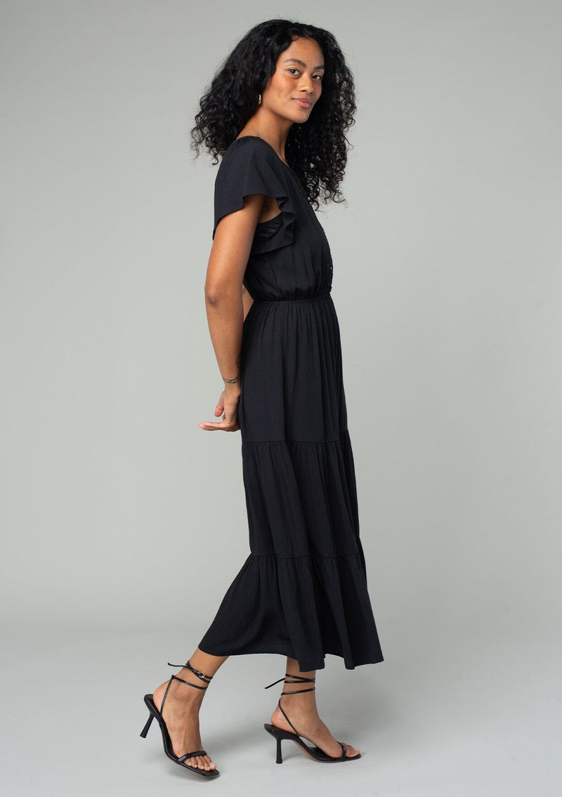 [Color: Black] A side facing image of a brunette model wearing a black bohemian spring mid length dress with short flutter sleeves, a tiered flowy skirt, an elastic waist, and lace trim detail at the top. 