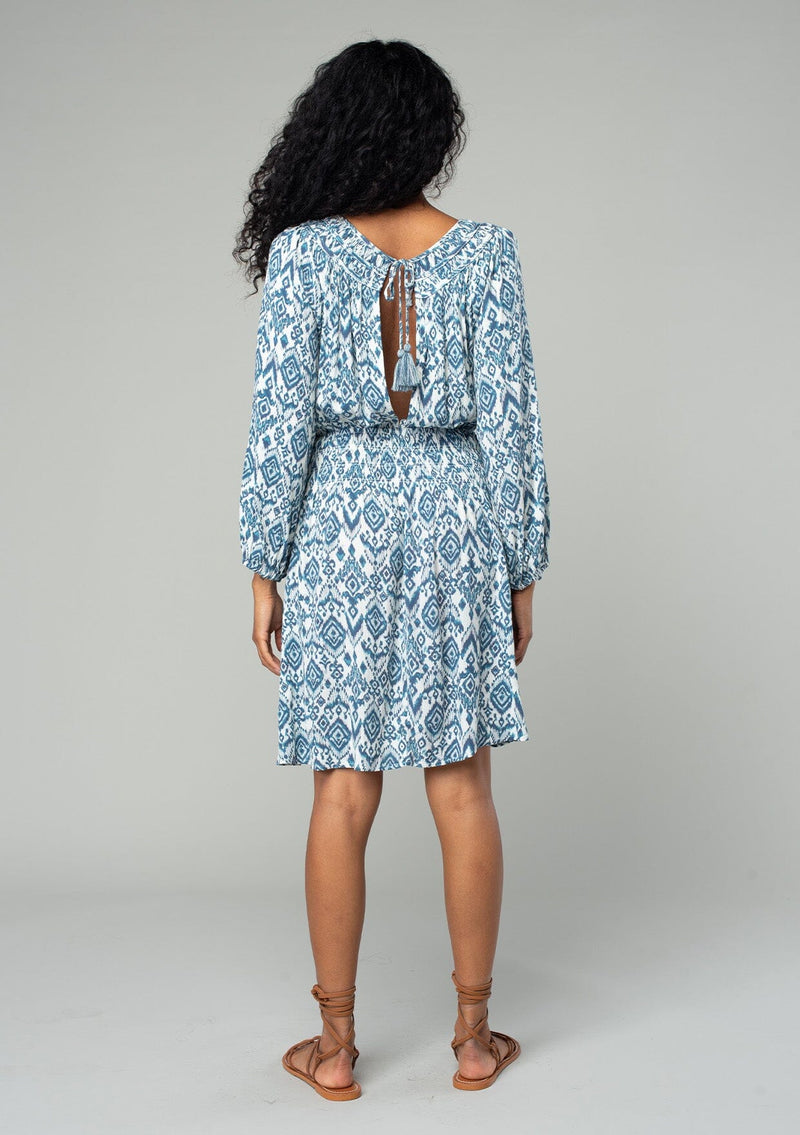 [Color: Ivory/Denim] A back facing image of a brunette model wearing a flowy spring mini dress in a bohemian blue and white abstract diamond print. With voluminous long sleeves, a smocked elastic waist, an open back with tassel tie closure, and a v neckline detail. 
