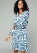 [Color: Ivory/Denim] A front facing image of a brunette model wearing a flowy spring mini dress in a bohemian blue and white abstract diamond print. With voluminous long sleeves, a smocked elastic waist, an open back with tassel tie closure, and a v neckline detail. 