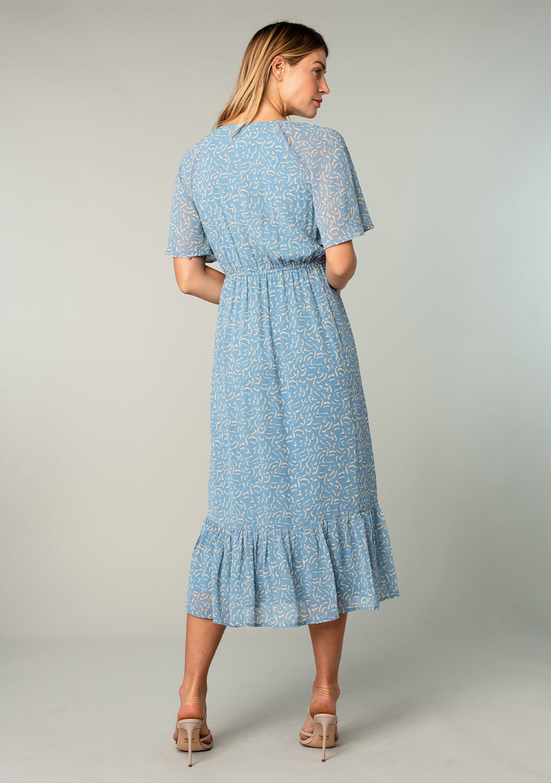 [Color: Dusty Blue/Natural] A back facing image of a blonde model wearing a bohemian blue mid length dress in lightweight sheer chiffon. With short flutter sleeves, a flattering top with gathered details, and a flowy tiered skirt. 