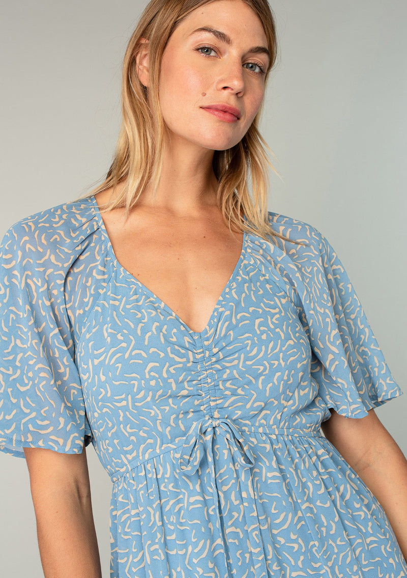 [Color: Dusty Blue/Natural] A close up front facing image of a blonde model wearing a bohemian blue mid length dress in lightweight sheer chiffon. With short flutter sleeves, a flattering top with gathered details, and a flowy tiered skirt. 