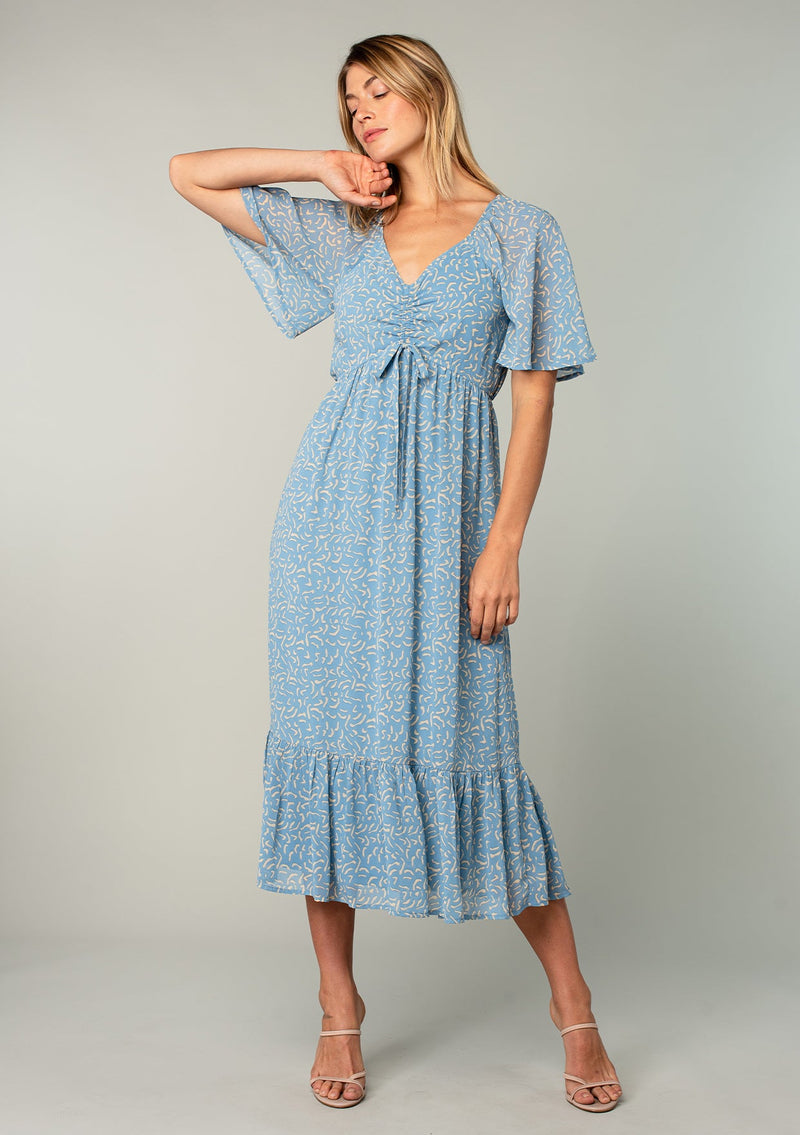 [Color: Dusty Blue/Natural] A front facing image of a blonde model wearing a bohemian blue mid length dress in lightweight sheer chiffon. With short flutter sleeves, a flattering top with gathered details, and a flowy tiered skirt. 
