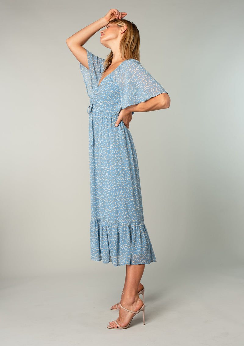 [Color: Dusty Blue/Natural] A side facing image of a blonde model wearing a bohemian blue mid length dress in lightweight sheer chiffon. With short flutter sleeves, a flattering top with gathered details, and a flowy tiered skirt. 