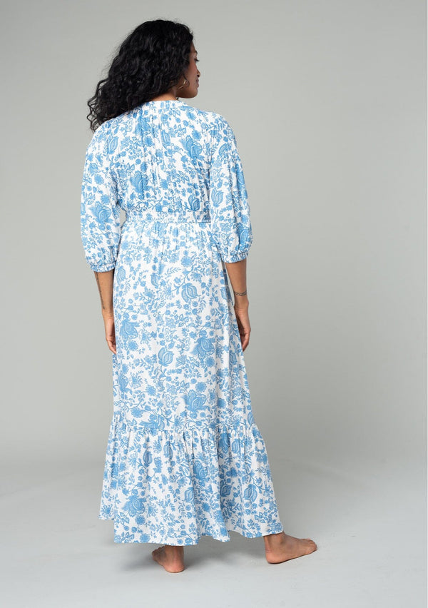 [Color: Cream/Dusty Blue] A back facing image of a brunette model wearing a bohemian cottage core style maxi dress in a white and blue floral print. With half length puff sleeves, a smocked elastic waist detail, a flowy tiered skirt, and a v neckline. 