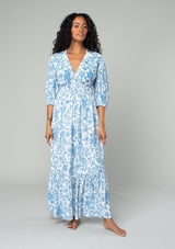 [Color: Cream/Dusty Blue] A front facing image of a brunette model wearing a bohemian cottage core style maxi dress in a white and blue floral print. With half length puff sleeves, a smocked elastic waist detail, a flowy tiered skirt, and a v neckline. 