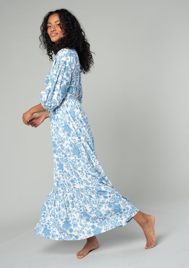 [Color: Cream/Dusty Blue] A side facing image of a brunette model wearing a bohemian cottage core style maxi dress in a white and blue floral print. With half length puff sleeves, a smocked elastic waist detail, a flowy tiered skirt, and a v neckline. 