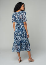 [Color: Indigo/Blue] A back facing image of a brunette model wearing a pretty bohemian mid length chiffon dress in a blue and navy blue floral print. With short sleeves, a self covered button front, a v neckline, and a back waist tie. 
