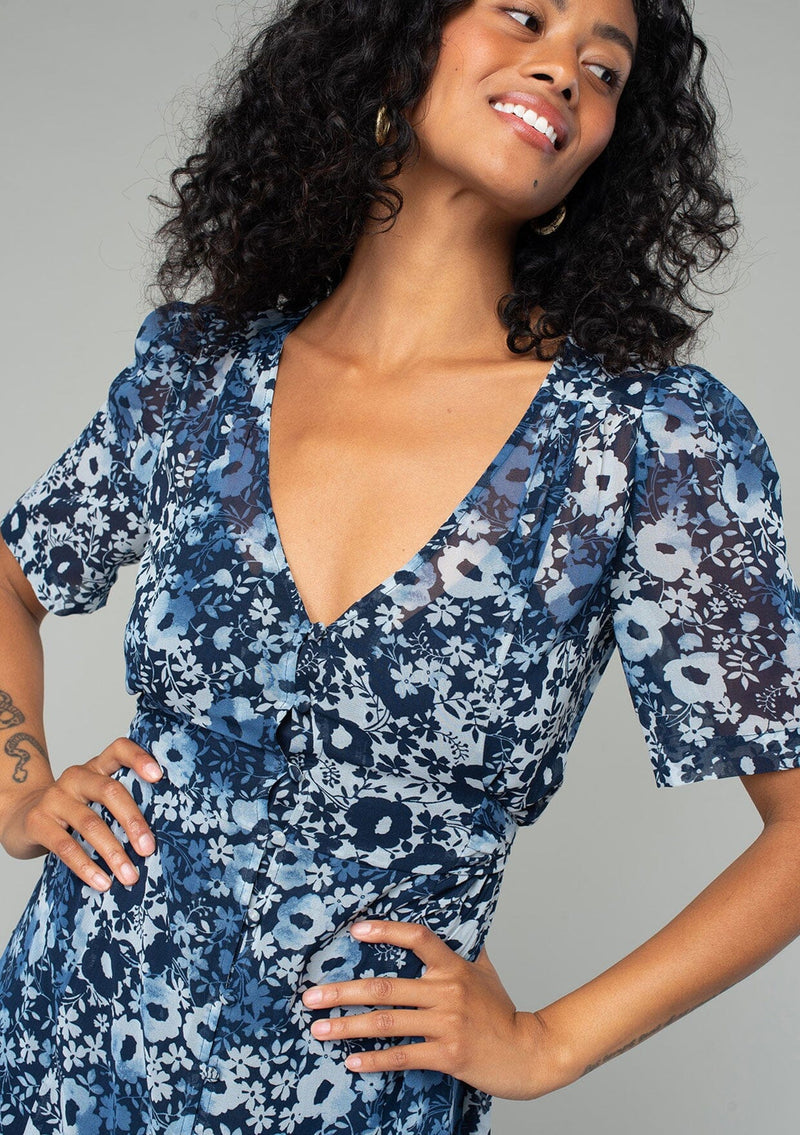 [Color: Indigo/Blue] A close up front facing image of a brunette model wearing a pretty bohemian mid length chiffon dress in a blue and navy blue floral print. With short sleeves, a self covered button front, a v neckline, and a back waist tie. 