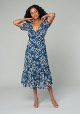 [Color: Indigo/Blue] A full body front facing image of a brunette model wearing a pretty bohemian mid length chiffon dress in a blue and navy blue floral print. With short sleeves, a self covered button front, a v neckline, and a back waist tie. 