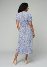 [Color: Blue/Coral] A back facing image of a brunette model wearing a bohemian spring mid length dress in a blue and coral floral print. With short puff sleeves, a button front, and side pockets. 