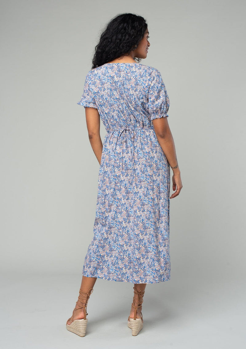 [Color: Blue/Coral] A back facing image of a brunette model wearing a bohemian spring mid length dress in a blue and coral floral print. With short puff sleeves, a button front, and side pockets. 