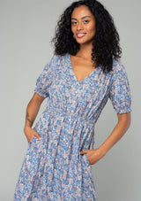 [Color: Blue/Coral] A close up front facing image of a brunette model wearing a bohemian spring mid length dress in a blue and coral floral print. With short puff sleeves, a button front, and side pockets. 