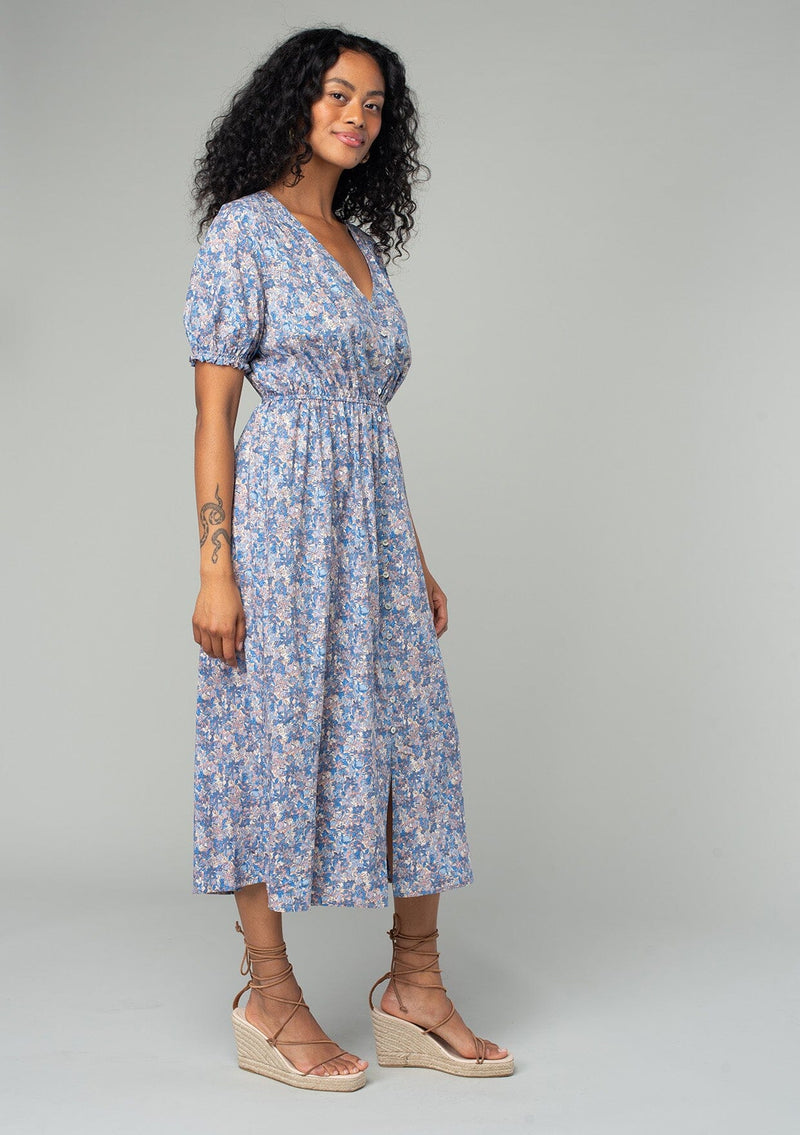 [Color: Blue/Coral] A side facing image of a brunette model wearing a bohemian spring mid length dress in a blue and coral floral print. With short puff sleeves, a button front, and side pockets. 