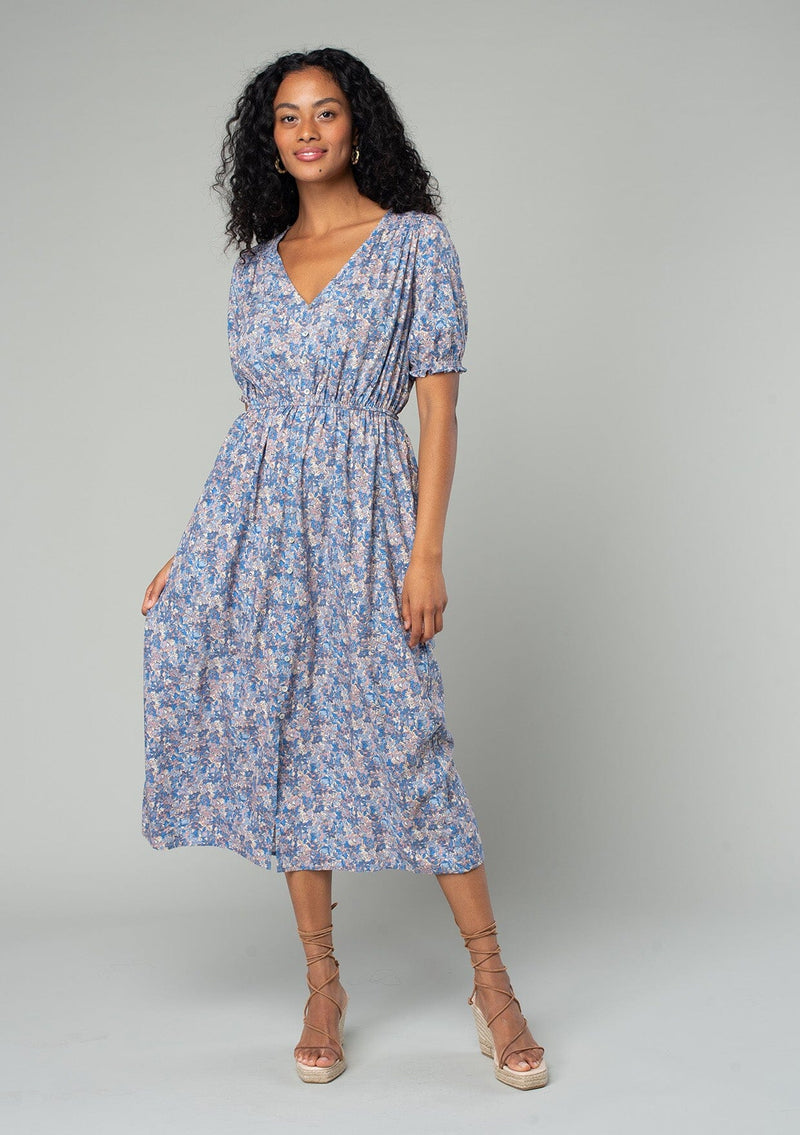 [Color: Blue/Coral] A front facing image of a brunette model wearing a bohemian spring mid length dress in a blue and coral floral print. With short puff sleeves, a button front, and side pockets. 