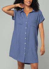 [Color: Dark Denim] A close up front facing image of a brunette model wearing a loose and relaxed fit blue mini shirt dress. With short cuffed sleeves, a button front, front patch pockets, and side pockets. 