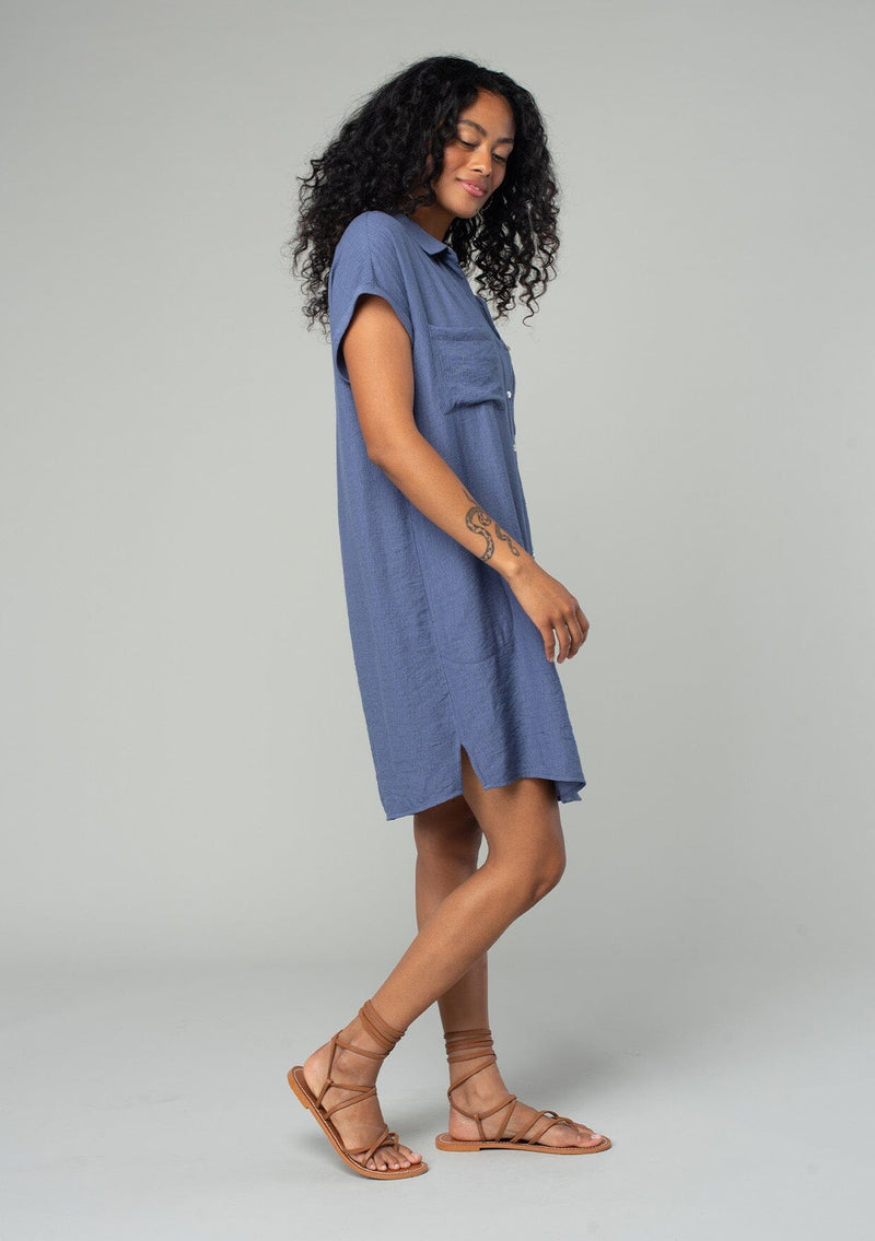 [Color: Dark Denim] A side facing image of a brunette model wearing a loose and relaxed fit blue mini shirt dress. With short cuffed sleeves, a button front, front patch pockets, and side pockets. 