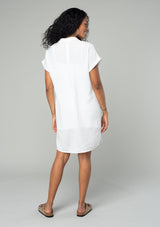 [Color: White] A back facing image of a brunette model wearing a loose and relaxed fit white mini shirt dress. With short cuffed sleeves, a button front, front patch pockets, and side pockets. 