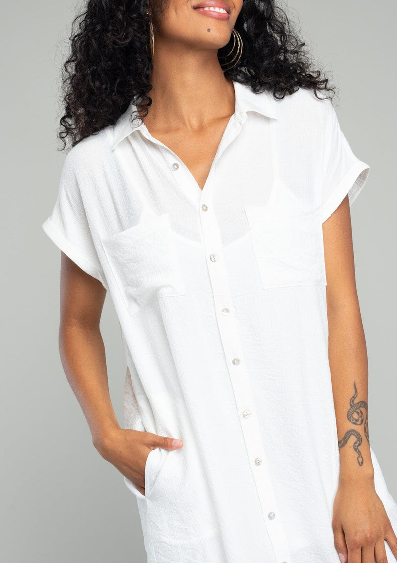 [Color: White] A close up front facing image of a brunette model wearing a loose and relaxed fit white mini shirt dress. With short cuffed sleeves, a button front, front patch pockets, and side pockets. 