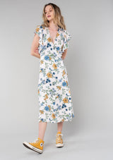 [Color: Natural/Mustard] A front facing image of a blonde model wearing a bohemian maxi wrap dress in a natural, blue, and mustard yellow floral print. A spring boho dress with short ruffled sleeves, a deep v neckline, and a side tie waist closure. 