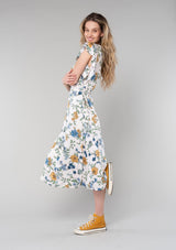 [Color: Natural/Mustard] A side facing image of a blonde model wearing a bohemian maxi wrap dress in a natural, blue, and mustard yellow floral print. A spring boho dress with short ruffled sleeves, a deep v neckline, and a side tie waist closure. 