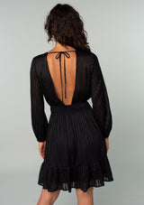[Color: Black] A back facing image of a brunette model wearing a bohemian holiday mini dress in a black lurex stripe. With long sleeves, a tiered flowy skirt, a smocked elastic waist, a v neckline, and an open back with tie closure.