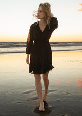 [Color: Black] A full body front facing image of a blonde model on the beach wearing a bohemian holiday mini dress in a black lurex stripe. With long sleeves, a tiered flowy skirt, a smocked elastic waist, a v neckline, and an open back with tie closure.